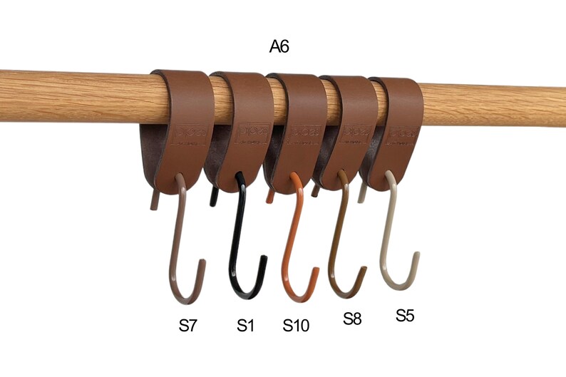 May include: Six brown leather hanging hooks with metal hooks in various colors, including black, orange, and beige, are attached to a wooden rod. The hooks are labeled with letters and numbers.