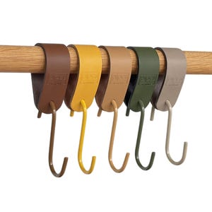 May include: Six leather and metal S-hooks in shades of brown, yellow, tan, green, and grey hang from a wooden rod. The hooks are designed for hanging items and have a minimalist aesthetic.