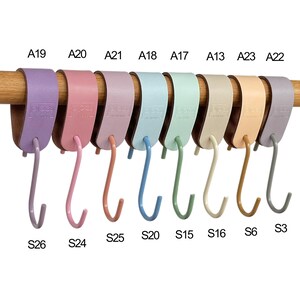 May include: A collection of colorful S-shaped hooks in various pastel shades, including lavender, pink, and blue, are displayed on a wooden rod. Each hook is labeled with a letter and number combination, such as A19 and S26.