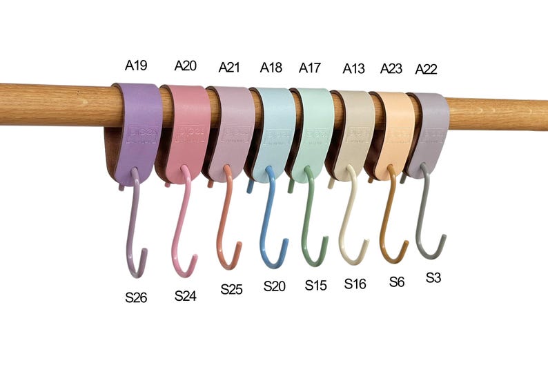 May include: A collection of colorful S-shaped hooks in various pastel shades, including lavender, pink, and blue, are displayed on a wooden rod. Each hook is labeled with a letter and number combination, such as A19 and S26.