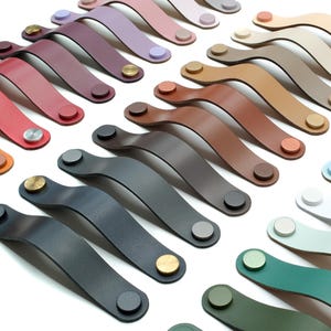 May include: A collection of curved leather drawer pulls arranged in rows, featuring a wide variety of solid colors including red, orange, yellow, blue, green, brown, black, white, and pastel shades, each secured with two metallic fasteners.