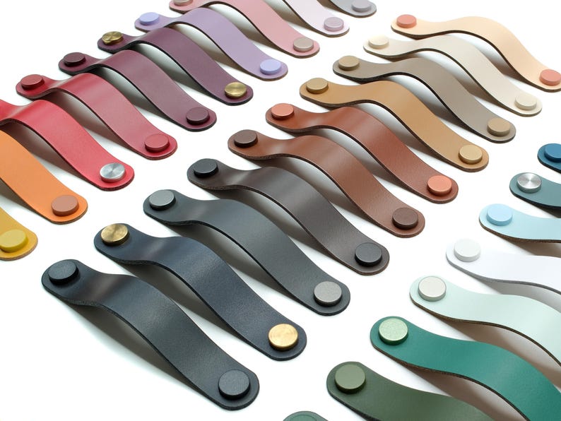 May include: A collection of curved leather drawer pulls arranged in rows, featuring a wide variety of solid colors including red, orange, yellow, blue, green, brown, black, white, and pastel shades, each secured with two metallic fasteners.