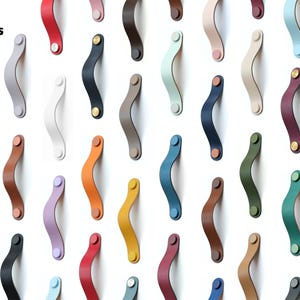 May include: A collection of Series ARC classic cabinet handles in various solid colors, including red, blue, yellow, green, black, white, and pastel shades. Each curved handle features two circular mounting points with metallic fasteners.