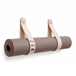 Puede incluir: Una esterilla de yoga marrón enrollada suspendida por dos correas de cuero beige claro fijadas a una pared. La correa izquierda presenta el texto Zoe Mia y un icono de flor de loto impresos en negro. Las correas se aseguran con cierres circulares.