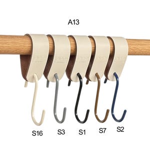May include: A row of six S-shaped hooks in various colors, suspended from a wooden rod. The hooks are cream, gray, black, brown, and navy blue. The hooks are attached to cream-colored leather straps.