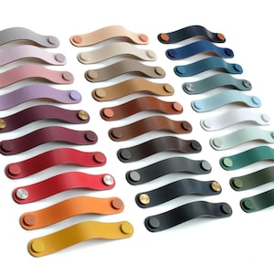 May include: A collection of arched leather drawer pulls arranged in three columns, featuring a wide variety of solid colors including shades of red, blue, green, yellow, brown, gray, and black, each secured with matching or metallic circular fasteners.