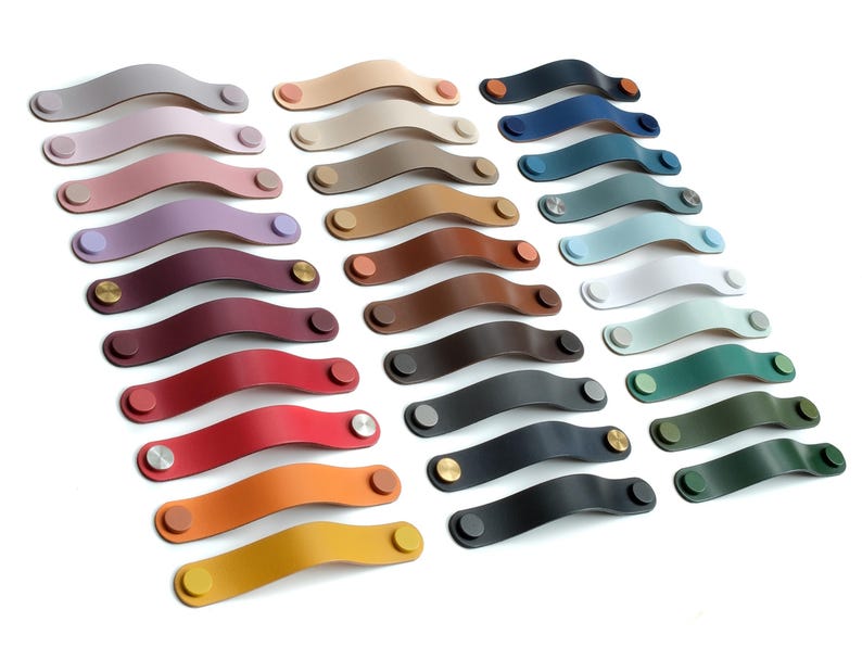 May include: A collection of arched leather drawer pulls arranged in three columns, featuring a wide variety of solid colors including shades of red, blue, green, yellow, brown, gray, and black, each secured with matching or metallic circular fasteners.