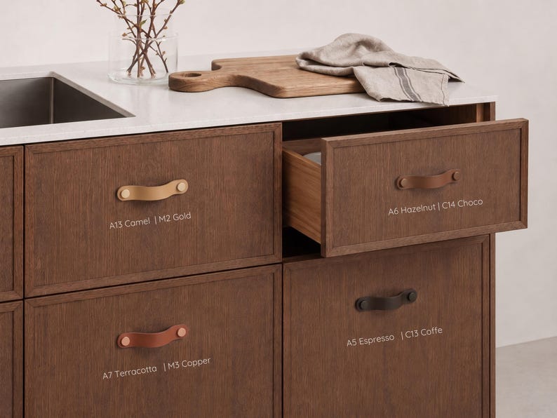 May include: Dark wood kitchen cabinetry featuring leather drawer pulls in various colors. Labels include A13 Camel | M2 Gold, A6 Hazelnut | C14 Choco, A7 Terracotta | M3 Copper, and A5 Espresso | C13 Coffe. One drawer is partially open, revealing the interior.