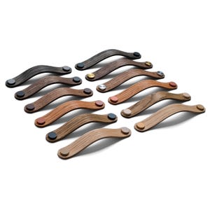 Puede incluir: Una colección de tiradores de madera para armarios en varios tonos de marrón, de oscuro a claro. Cada tirador tiene un diseño curvo y está fijado con un pequeño acento metálico circular en colores como plata, oro y cobre. Los tiradores están sobre un fondo blanco.