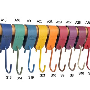 May include: A collection of colorful, leather-wrapped hooks hanging from a wooden rod. The hooks are in various colors, including blue, green, yellow, orange, red, and purple. Each hook has a metal hook attached. The hooks are labeled with letters and numbers.