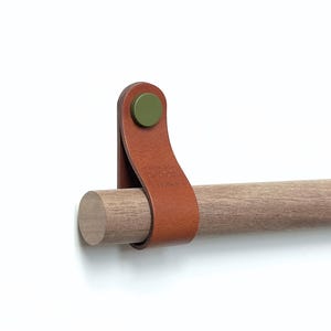 Può includere: Un'asta di legno con un cinturino in pelle marrone e un bottone verde. L'asta è fissata a una parete con una vite.