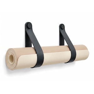Può includere: Un supporto da parete composto da due cinghie in pelle nera fissate con elementi circolari neri, che sostiene un tappetino da yoga color beige chiaro arrotolato in posizione orizzontale su uno sfondo bianco.