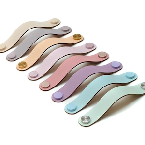 Op de afbeelding: Een collectie van acht gebogen ladehandgrepen van leer in diverse pastelkleuren, waaronder crème, grijs, beige, roze, lavendel en lichtblauw. Elke handgreep is aan beide uiteinden bevestigd met ronde metalen sluitingen in goud- of zilverkleur.