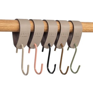 Könnte beinhalten: Ein Set aus sechs dekorativen Haken in verschiedenen Farben, die an einer Holzstange hängen. Die Haken sind in Beige, Rosa, Schwarz, Braun und Grün gehalten. Die Haken sind an einem beigefarbenen Lederriemen befestigt.