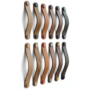 May include: A collection of wooden cabinet pulls in various shades of brown, from light oak to dark walnut. Each pull has a curved, ergonomic design and is attached with a contrasting colored metal button. The pulls are arranged in two rows against a white background.