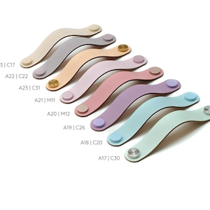 May include: A collection of eight curved leather drawer pulls in various pastel and neutral colors, including beige, gray, tan, pink, lavender, blue, and mint. Each pull features two circular metal or matching leather fasteners at the ends.