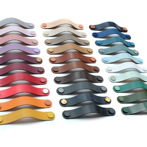 Puede incluir: Una colección de tiradores de cajón de cuero arqueados dispuestos en tres columnas. Los tiradores vienen en una variedad de colores, incluyendo tonos de rojo, azul, verde, amarillo, marrón, gris y blanco, asegurados con cierres metálicos circulares.