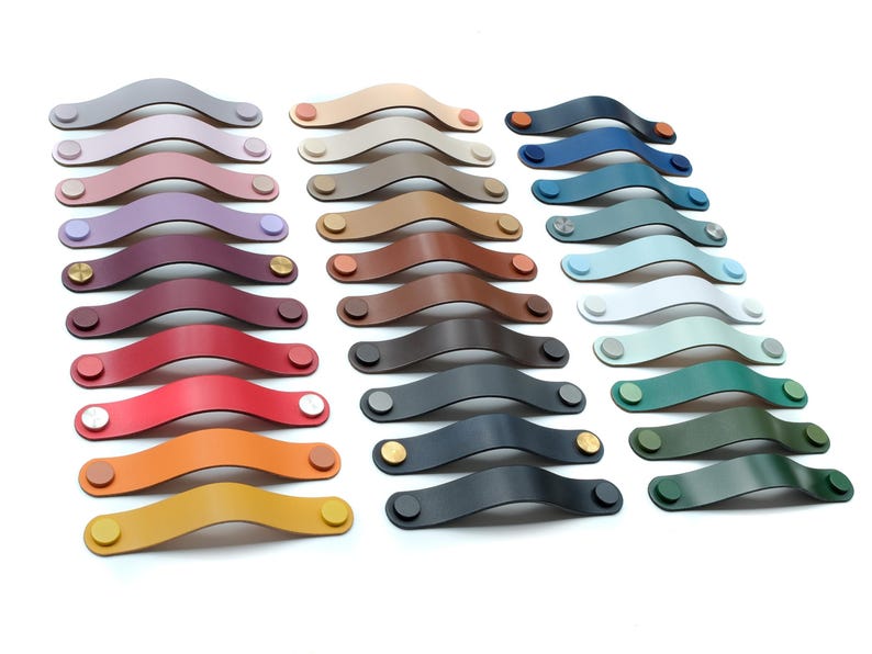 May include: A collection of arched leather drawer pulls arranged in three columns. The pulls come in a variety of colors including shades of red, blue, green, yellow, brown, grey, and white, each secured with circular metal fasteners at both ends.