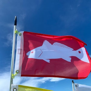 Könnte beinhalten: Eine rote Flagge mit einer weißen Silhouette eines Fisches, mit einem roten Auge, die im Wind gegen einen blauen Himmel weht.
