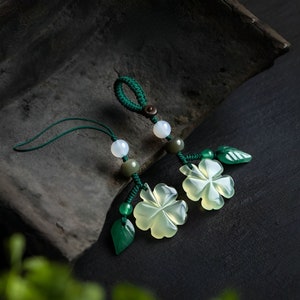 Puede incluir: Dos amuletos de trébol de jade verde con detalles de hojas verdes en una cuerda verde con cuentas blancas y verdes. Los amuletos cuelgan de una superficie gris oscuro.