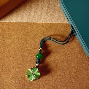 Puede incluir: Un charm de teléfono verde con un colgante de trébol de cuatro hojas. El charm presenta un cordón verde, una cuenta dorada y una cuenta verde. El colgante de trébol tiene un borde dorado y un centro verde translúcido. La funda del teléfono es verde azulado.