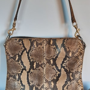 Könnte beinhalten: Eine rechteckige Handtasche mit Schlangenmuster in Braun-, Beige- und Schwarztönen. Die Tasche hat einen langen, dünnen Riemen und silberfarbene Beschläge. Die Tasche ist eine Umhängetasche.