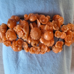 May include: A coral-colored bracelet featuring carved floral and fruit designs. The bracelet is composed of various shapes, including flowers, spheres, and a central sunflower. The intricate details and warm color create a vintage aesthetic. The bracelet is displayed on a blue fabric.