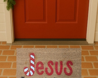 May include: A rectangular doormat with the word "Jesus" in red, with a candy cane design. Below, the text reads "The reason for the season" in black. The mat is placed in front of a red door.