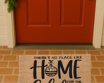 May include: A rectangular doormat with the text "THERE'S NO PLACE LIKE HOME for the holidays" in black script. The mat is beige and sits in front of a red door. A Christmas wreath with red berries hangs on the left.