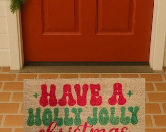 May include: A rectangular doormat with the words "HAVE A HOLLY JOLLY christmas" in red and green lettering. The mat is beige and sits in front of a red door. A green wreath with red berries is above the door.