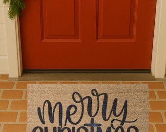 May include: A rectangular doormat with the words "merry CHRISTMAS" in a dark blue script. The mat is beige and sits on a brick surface in front of a red door. A green garland with red berries is above the door.