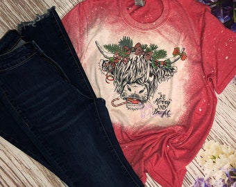 May include: Red bleached t-shirt with a white design of a Highland cow wearing a Christmas wreath and candy cane. The text "Be Merry and Bright" is below the cow.