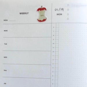 Peut inclure: Une page d'agenda hebdomadaire avec le mot "WEEKLY" en haut. Un autocollant de trognon de pomme est plac&eacute; au-dessus des lignes de l'agenda. La date "01/19" et le jour "MON" sont &eacute;crits sur la page. La page a une grille pointill&eacute;e.