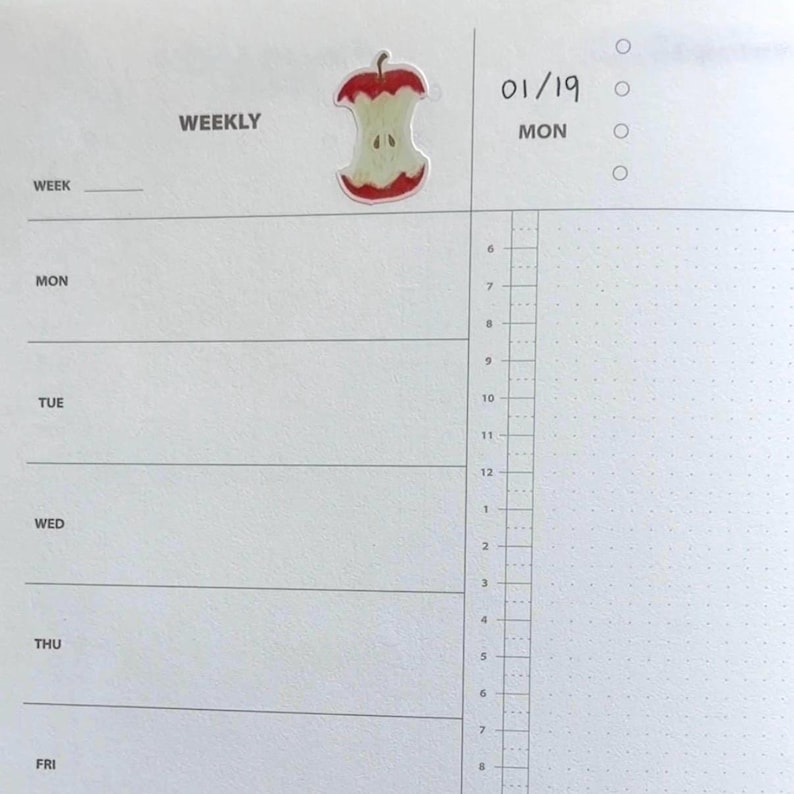 Peut inclure: Une page d'agenda hebdomadaire avec le mot "WEEKLY" en haut. Un autocollant de trognon de pomme est plac&eacute; au-dessus des lignes de l'agenda. La date "01/19" et le jour "MON" sont &eacute;crits sur la page. La page a une grille pointill&eacute;e.