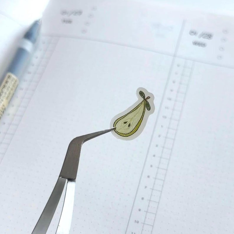 Peut inclure: Un autocollant en forme de poire avec un design vert et jaune est tenu par une pince &agrave; &eacute;piler argent&eacute;e. L'autocollant a un contour d&eacute;taill&eacute; et une petite feuille verte en haut. Le fond est un cahier blanc avec un motif quadrill&eacute;.