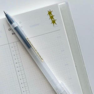 Peut inclure: Un stylo &agrave; bille &agrave; encre gel transparente avec de l'encre noire, pos&eacute; sur un cahier blanc &agrave; points. Le stylo a un capuchon transparent et une &eacute;tiquette avec du texte noir. Quelques autocollants jaunes en forme d'&eacute;toile sont sur le cahier.