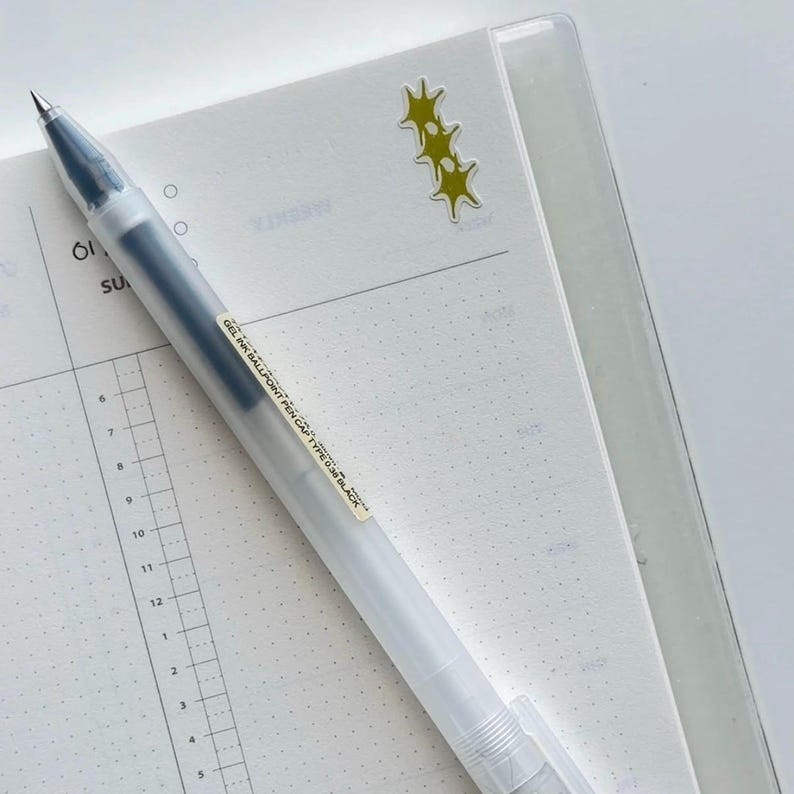 Peut inclure: Un stylo &agrave; bille &agrave; encre gel transparente avec de l'encre noire, pos&eacute; sur un cahier blanc &agrave; points. Le stylo a un capuchon transparent et une &eacute;tiquette avec du texte noir. Quelques autocollants jaunes en forme d'&eacute;toile sont sur le cahier.