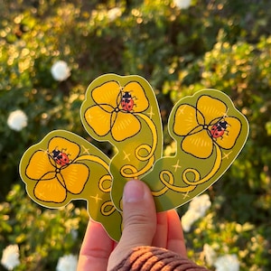 May include: Three stickers featuring yellow flowers with a ladybug in the center. The stickers have a green background with a gold swirl design. The stickers are held in a person's hand, with a blurred background of greenery.