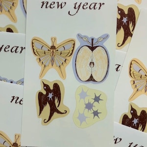 Puede incluir: Una colección de pegatinas con temática de año nuevo. Las pegatinas presentan ilustraciones de mariposas, manzanas, pájaros y estrellas en tonos amarillos, marrones y azules. Las palabras "new year" están impresas en algunas pegatinas.