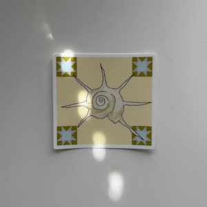 May include: Square sticker with a beige background featuring a black line drawing of a spiral shell with radiating lines. The corners have a blue and gold geometric design. The sticker is on a white surface.
