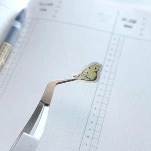 Peut inclure: Un petit autocollant d'oiseau jaune clair tenu par une pince &agrave; &eacute;piler argent&eacute;e. L'autocollant a un contour d&eacute;taill&eacute; et est positionn&eacute; sur une page de carnet blanche lign&eacute;e. Un stylo bleu est visible en arri&egrave;re-plan.