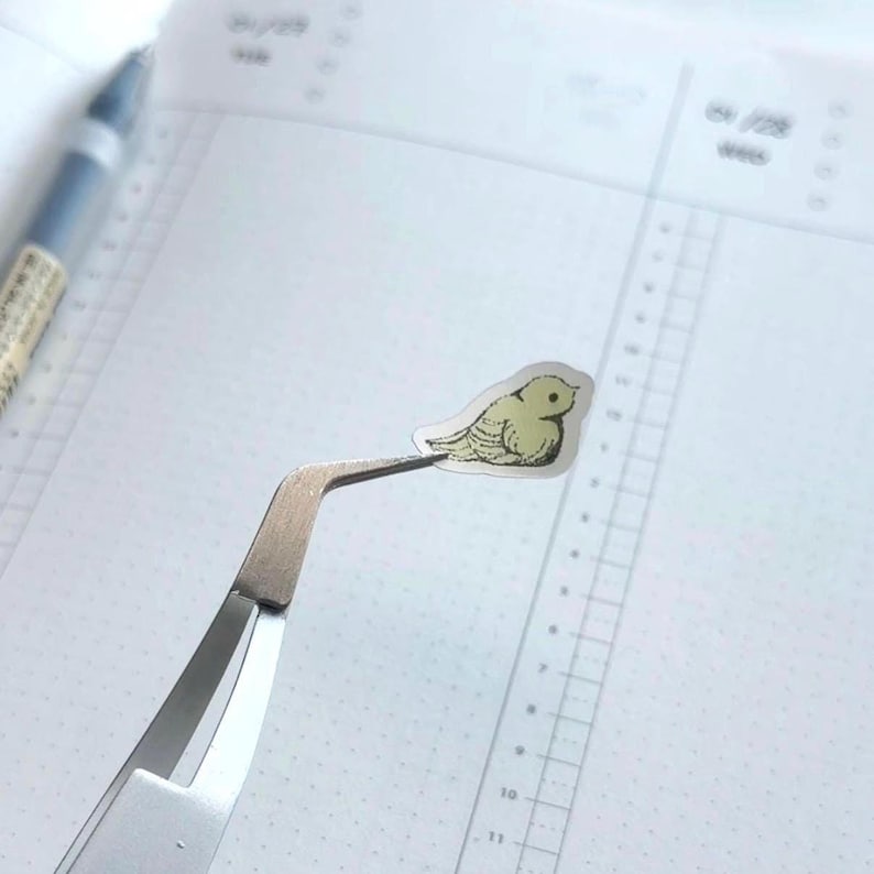Peut inclure: Un petit autocollant d'oiseau jaune clair tenu par une pince &agrave; &eacute;piler argent&eacute;e. L'autocollant a un contour d&eacute;taill&eacute; et est positionn&eacute; sur une page de carnet blanche lign&eacute;e. Un stylo bleu est visible en arri&egrave;re-plan.