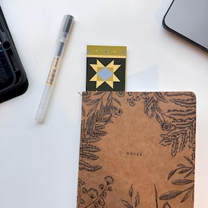May include: A brown notebook with the word "NOTES" printed on the cover. A decorative bookmark with a geometric design is inserted into the notebook. A clear pen and a black phone case are also visible.