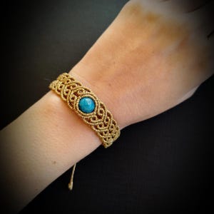 May include: A handmade macrame bracelet with a central turquoise stone. The bracelet is crafted with tan-colored cord, featuring intricate knotting patterns. The round turquoise stone is the focal point of the bracelet.