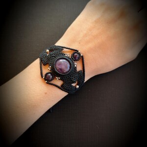 Peut inclure: Bracelet en macramé noir orné d'une grosse pierre précieuse ronde et violette et de petites pierres précieuses violettes. Le bracelet a un design carré avec des accents argentés. Le bracelet est porté au poignet.