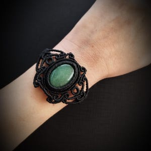 Puede incluir: Un brazalete de macramé negro con una piedra preciosa verde ovalada. La piedra está engastada en un marco decorativo calado. El brazalete se lleva en la muñeca.