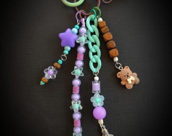 May include: A decorative keychain with multiple charms. The keychain features a variety of beads, including purple stars, flowers, and a small brown teddy bear. The color palette includes shades of purple, green, and brown.