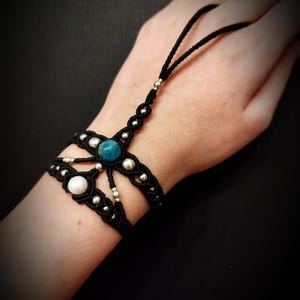 Puede incluir: Pulsera de mano de macramé negro con una gema azul y una perla blanca. La pulsera tiene cuentas plateadas y un lazo para el dedo. La joya está hecha a mano y es adecuada para diversas ocasiones.