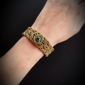 Peut inclure: Bracelet en macramé beige et marron, avec un motif tissé complexe. Le bracelet comporte une pierre centrale, ronde, vert foncé. Le bracelet est porté sur un poignet sur fond sombre.