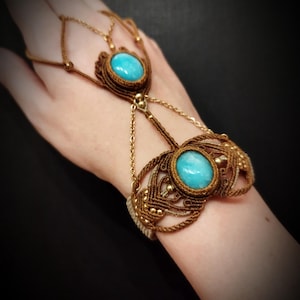 Könnte beinhalten: Ein handgefertigtes Makramee-Handkettenarmband mit zwei ovalen türkisfarbenen Steinen. Das Armband ist aus braunem Kordel und goldfarbener Kette und Perlen gefertigt und kreiert einen Bohemian-Stil.